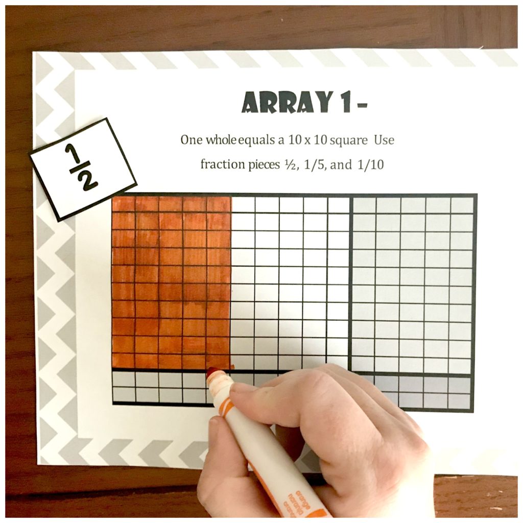 Multiplication arrays game worksheet on a wooden table with a child's hand coloring in the squares. 