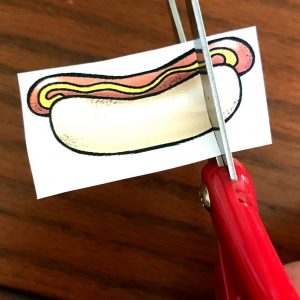Image of a hotdog being cut with red scissors. 
