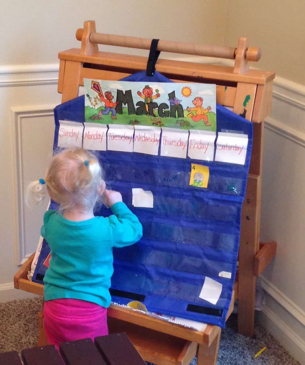 Pocket chart calendar with a little girl placing a calendar piece into a pocket. 