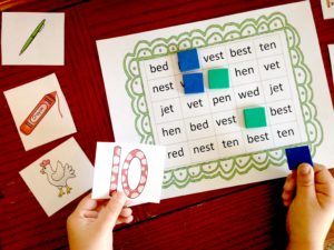 A short e game printable with game pieces and a little child's hands holding a card that says, 10 and a blue block in another hand. 