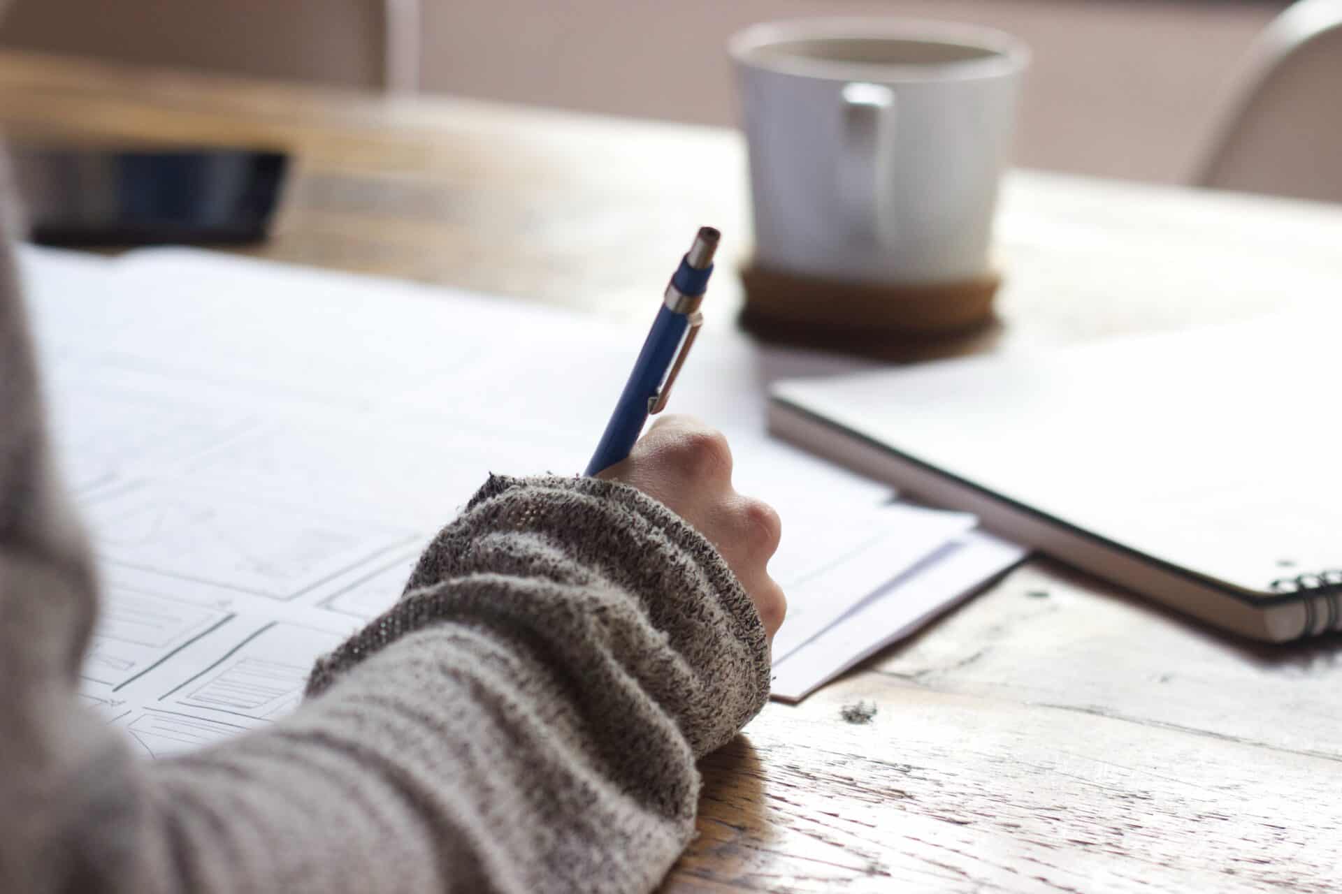 A teacher's hand holding a pen is grading work on a wooden desk. 